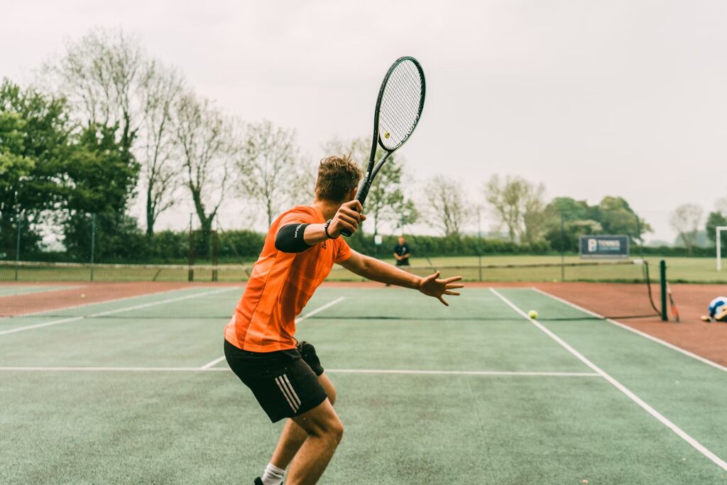 Jogador de tênis com uniforme laranja com detalhes verdes, com a raquete levantada para rebater uma bola.