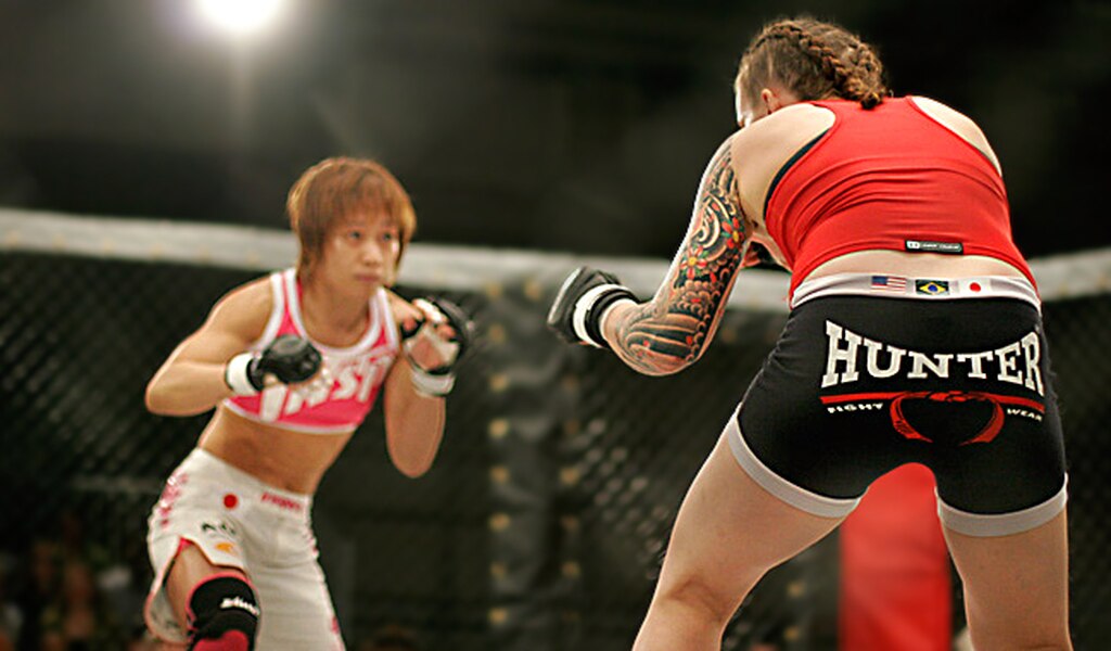 Luta do UFC feminino, com uma lutadora sendo vista de costas, enquanto que a outra, vestindo calção branco, é vista de frente.