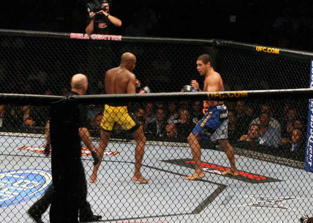 Luta do UFC sendo transmitida ao vivo, com lutador de calção amarelo indo para cima do de calção azul.