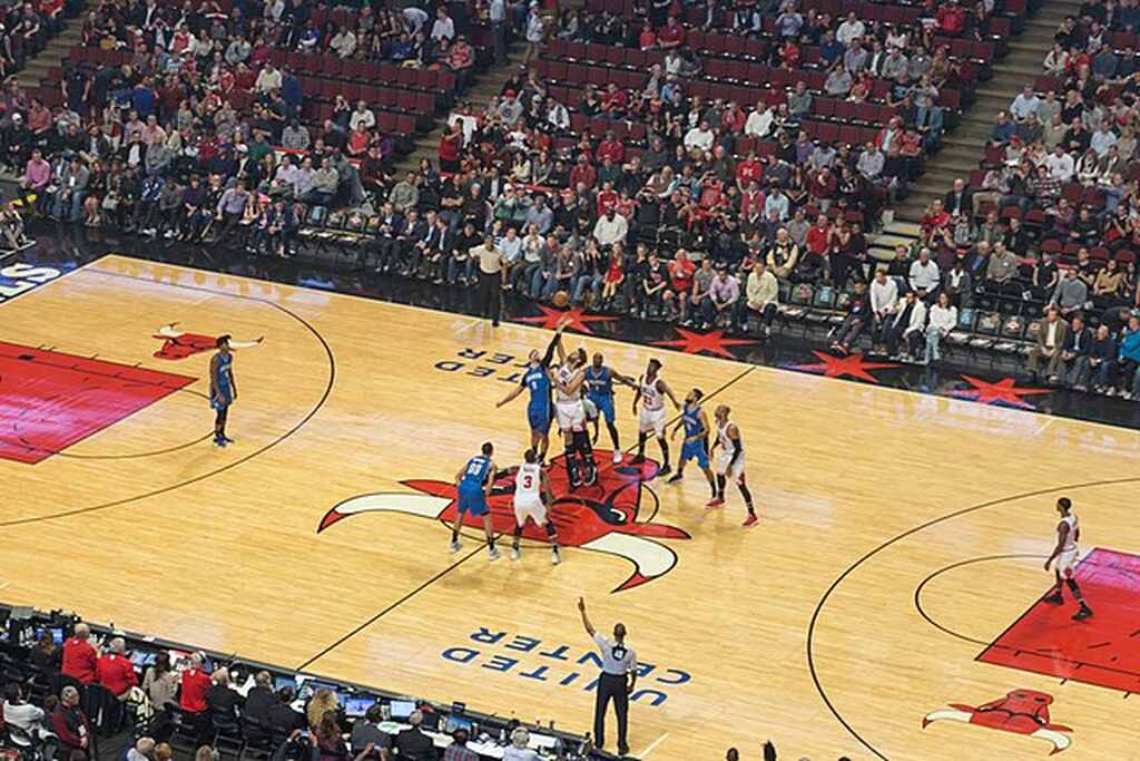 Início de partida de basquete da NBA, com jogador do time branco disputando bola no ar com jogador de time azul.