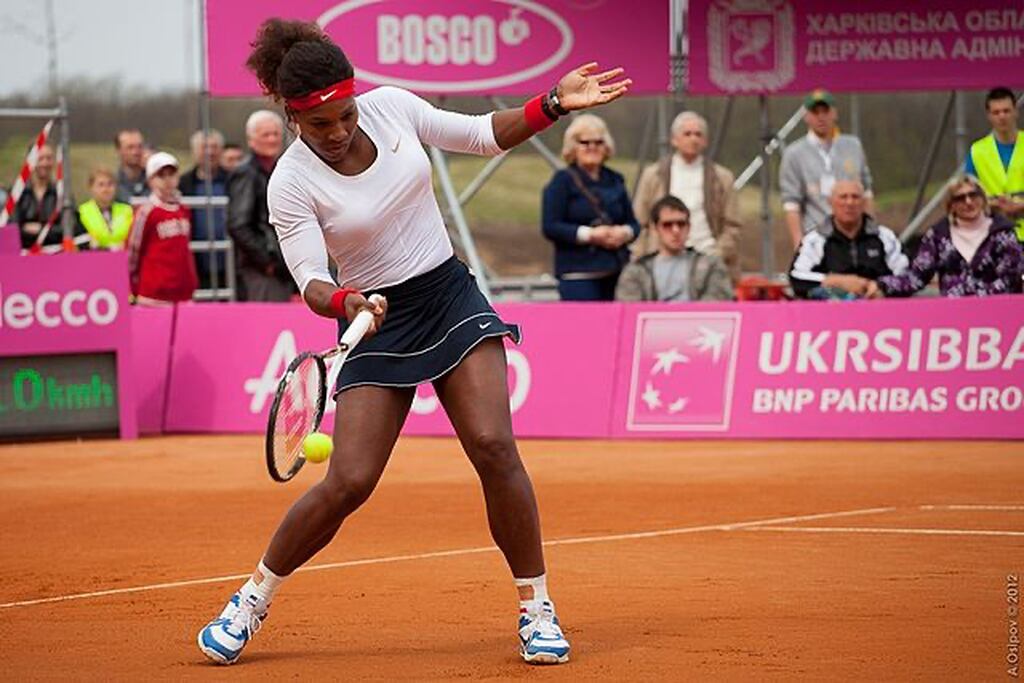 Ex-jogadora de tênis, vestindo roupa branca com short preto, Serena Williams, rebatendo uma bola durante partida.