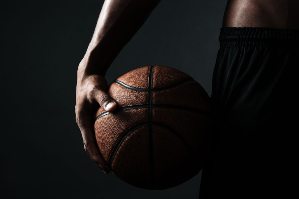 Jogador de basquete segurando uma bola laranja com a mão direita, encostada em sua cintura.