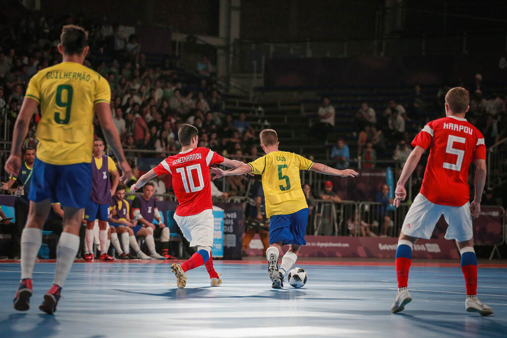 Jogador da seleção brasileira de futsal conduzindo a bola enquanto adversário de vermelho tenta desarmar.