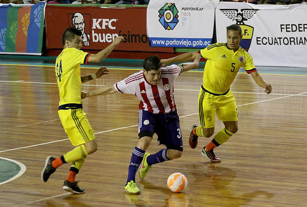 Jogador de Futsal vestindo camisa listrada passando por dois jogadores de uniforme amarelo.