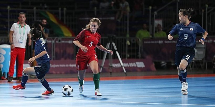 Jogadora de futsal vestindo uniforme vermelho, conduzindo a bola pela quadra, cercada por adversárias.