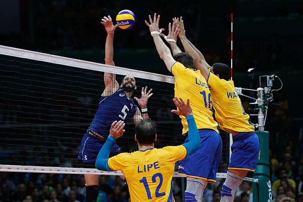 Seleção brasileira de vôlei masculino durante partida. Há dois atletas defendendo a bola por cima da rede.