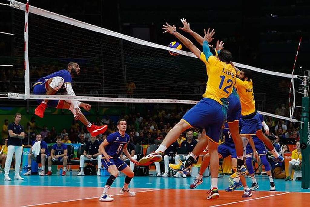 Seleção brasileira de vôlei masculino durante a partida. Há três atletas defendendo a bola por cima da rede.