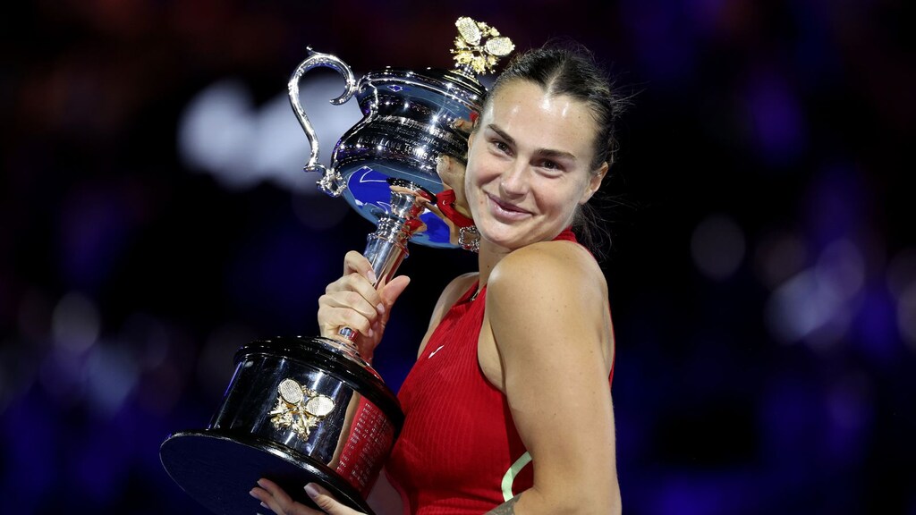 Jogadora de tênis Aryna Sabalenka, vestindo roupa vermelha, sorrindo e erguendo grande troféu.