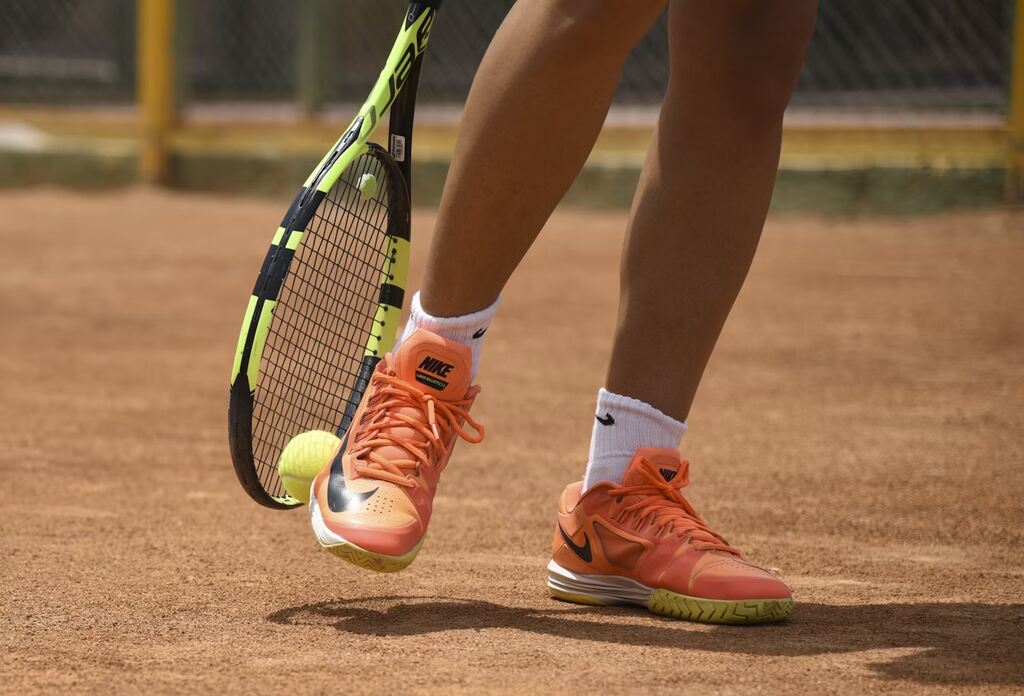 Jogadora de tênis segurando raquete com bolinha próxima do tênis em quadra de terra.