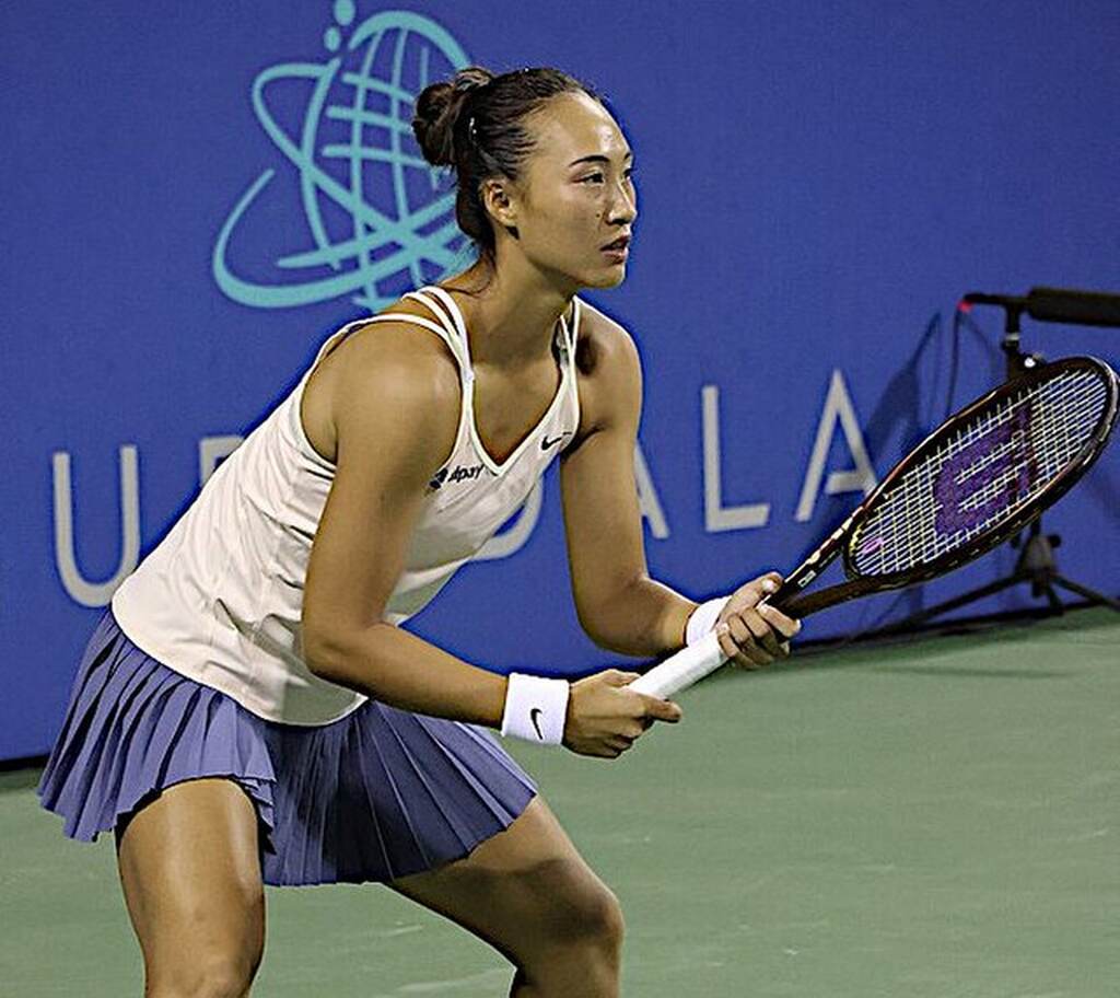 Jogadora de tênis Zheng Qinwen, vestindo uniforme branco com saia azul, segurando a raquete com as duas mãos.